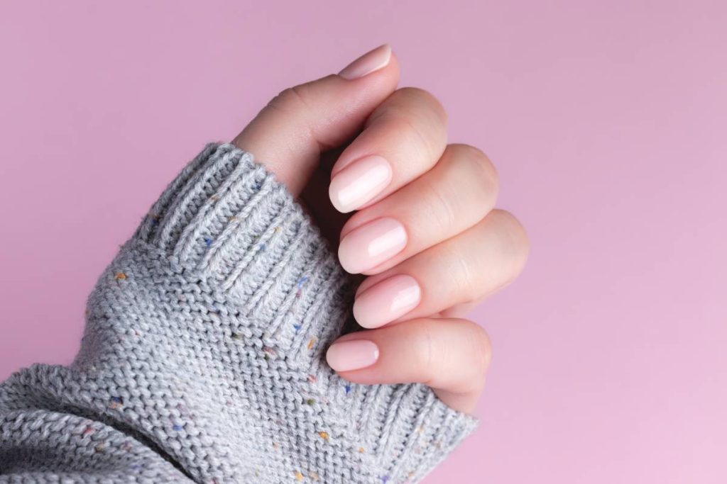 Female hand in gray knitted sweater with natural beautiful manicure - pink nude nails on pink background. Nail care concept